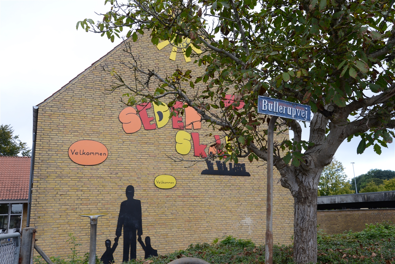 "Murstensbygning med farverigt skilt 'Seden Skole' og 'Velkommen', set bag et træ og et vejskilt med navnet 'Bullerupvej'."