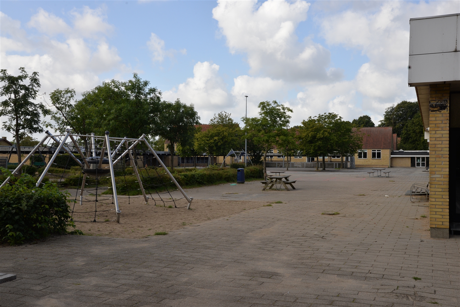 En legeplads med grønne træer og buske, terrassesten, bænkebord, en skældespand, en flagstang samt et edderkop legestativ. Der er murstensbygninger i baggrunden. 