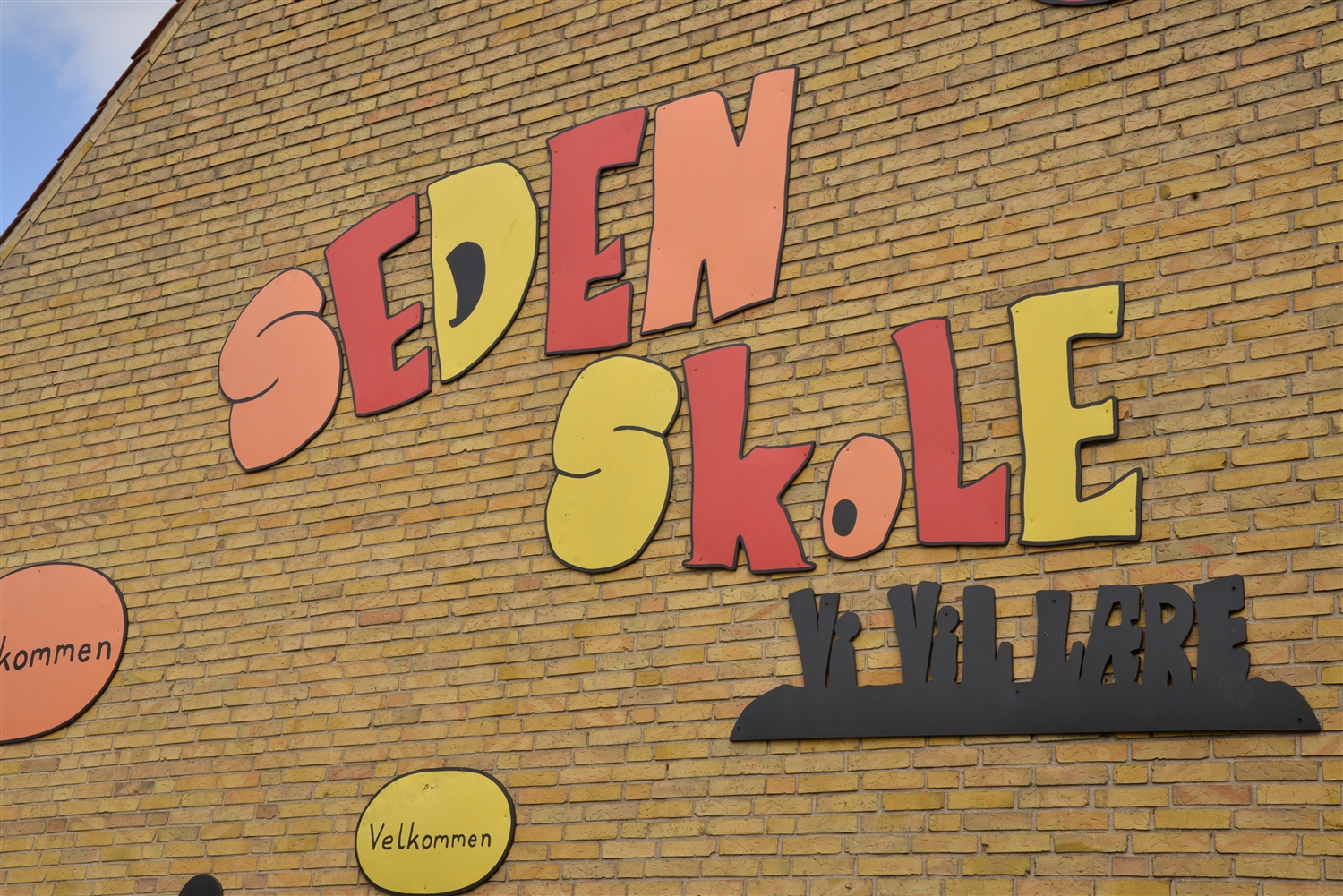 Murstensbygning med farverigt skilte "Seden skole" og "velkommen" samt et sort skilt "Vi vil lære".