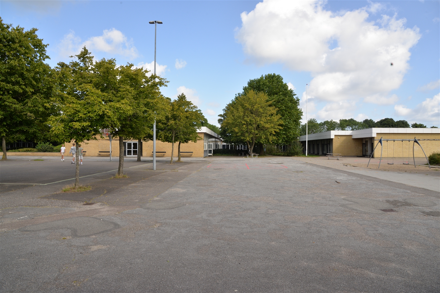 Murstensbygning i baggrunden. Grønne træer og buske foran, en flagstang og gyngestativ til venstre og en lygtepæl og et par børn der leger til højre. 