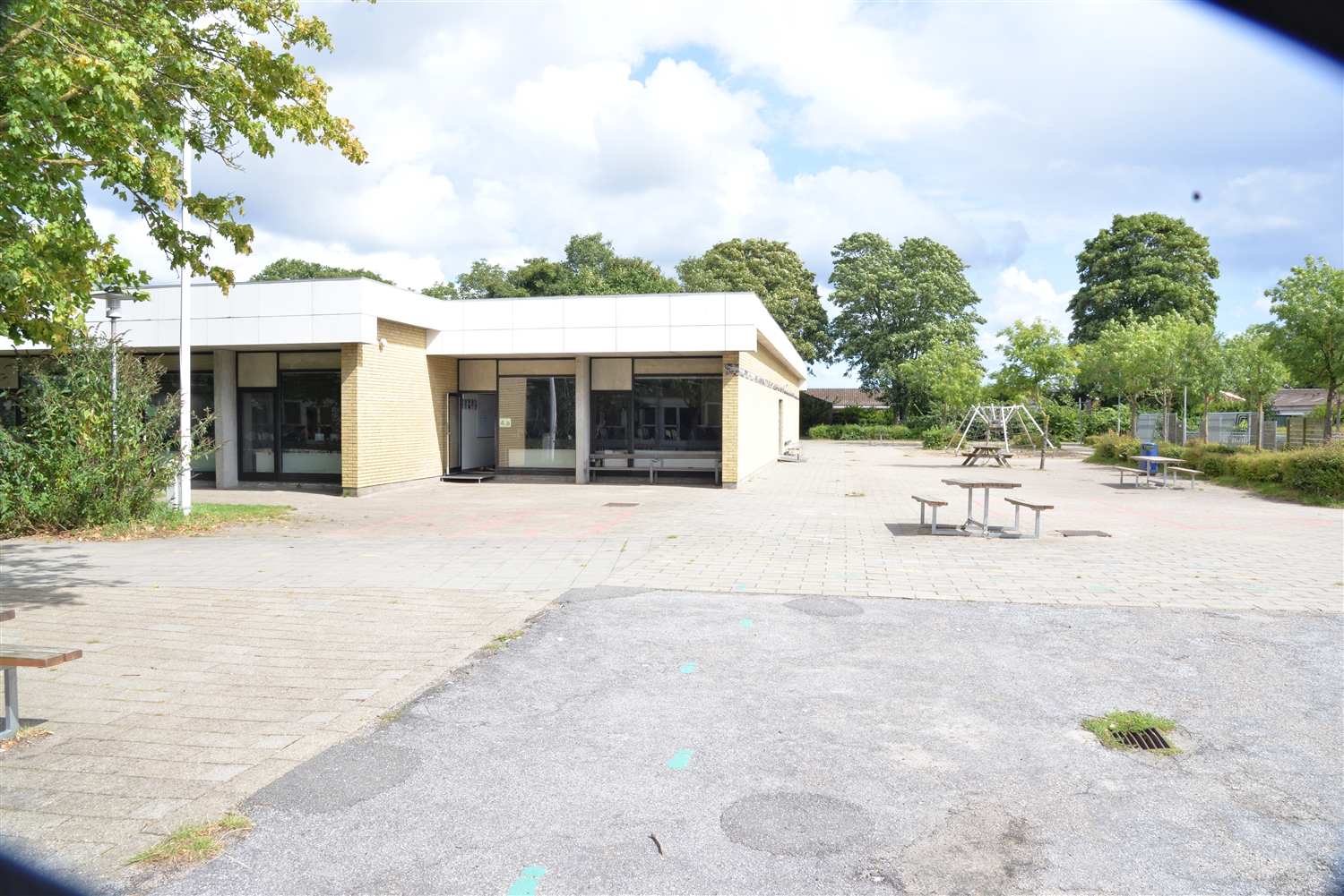 Grå gårdsplads med en murstensbygning. Til højre ses grønne træer og buske samt en flagstang. Til venstre ses grønne træer og buske samt et bænkebord og et edderkop legestativ.