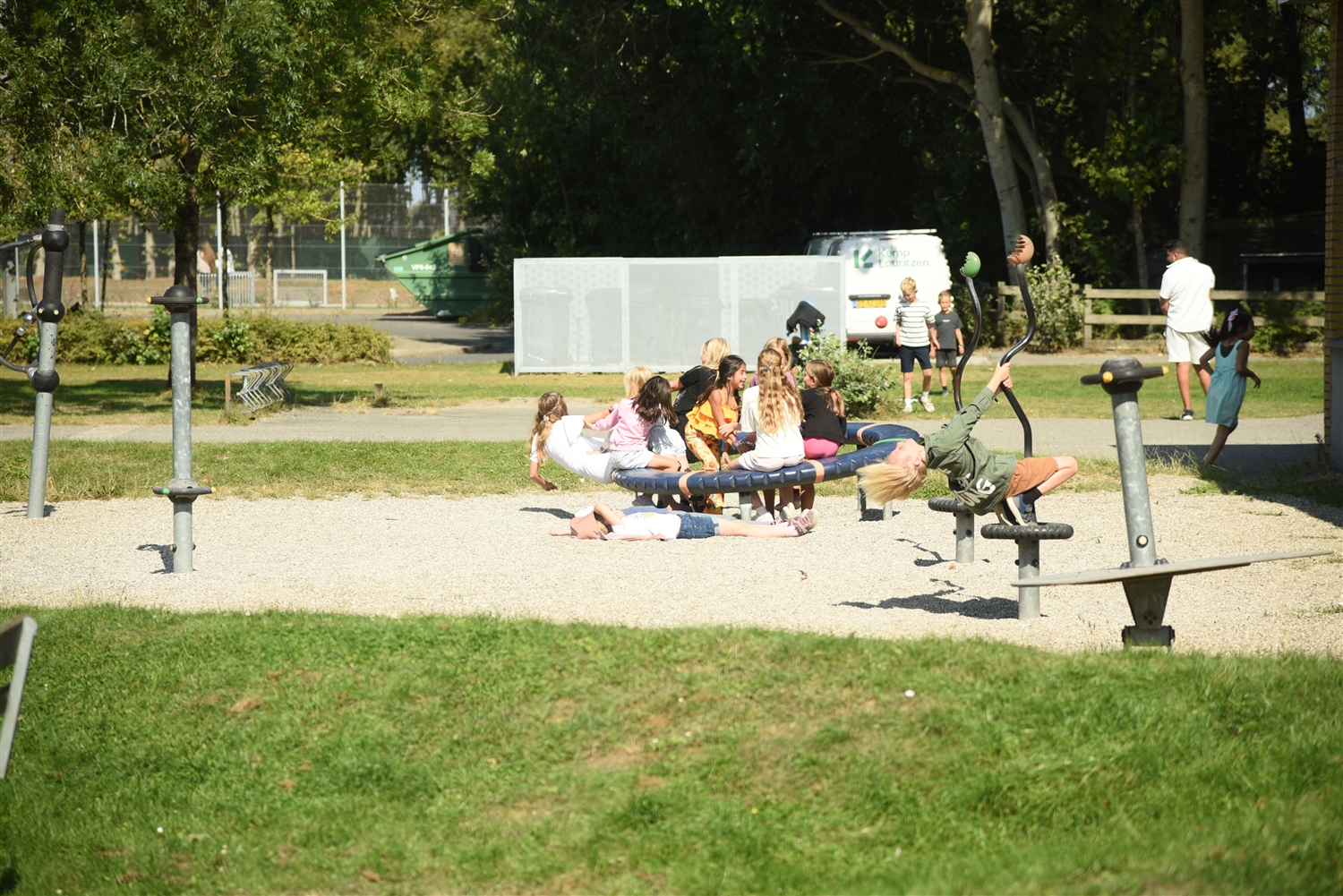 Sandlegeplads med omliggende græs. Træer og buske ses i bagrungen samt hvidt stativ og en varevogn. På sandet sidder der børn og nyder solskinnet. 
