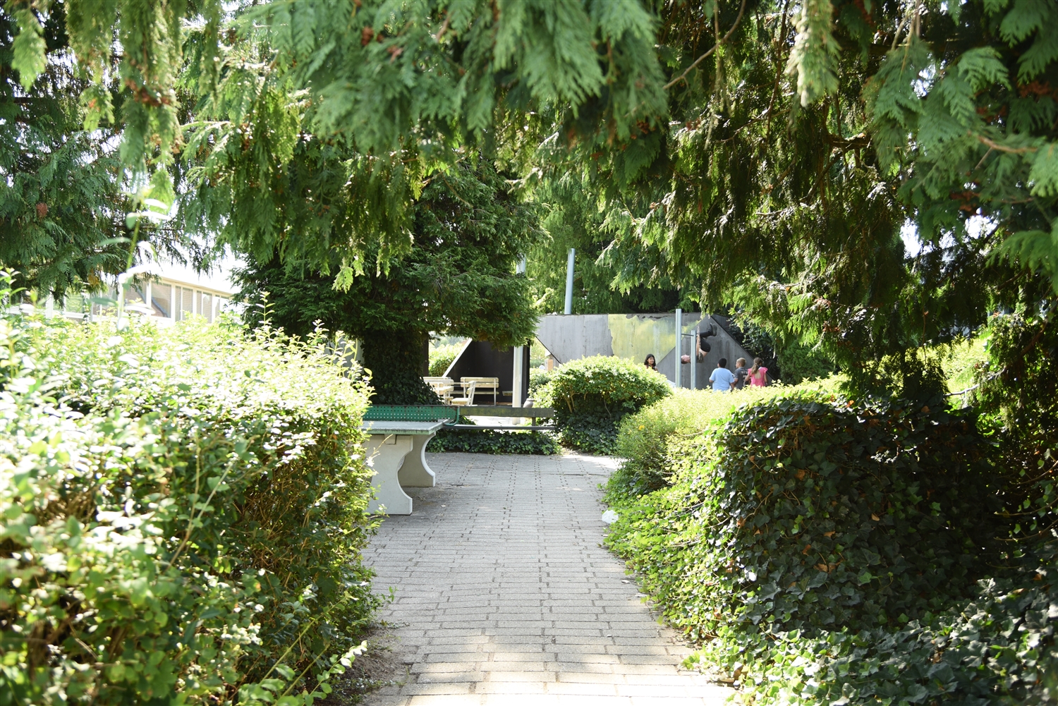 En gangsti omgivet af træer og buske. Til højre kan man se halvdelen af et bordtennisbord. Til venstre i bagrunden kan man se børn der leger. 