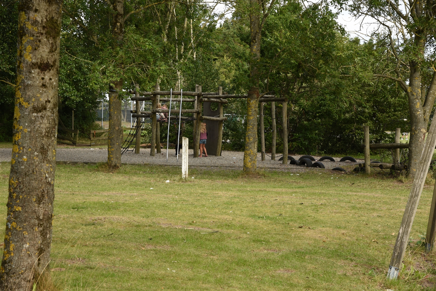 En klatrelegeplads på sand omgivet af græs, træer og buske. På legepladsen ses børn der leger. 