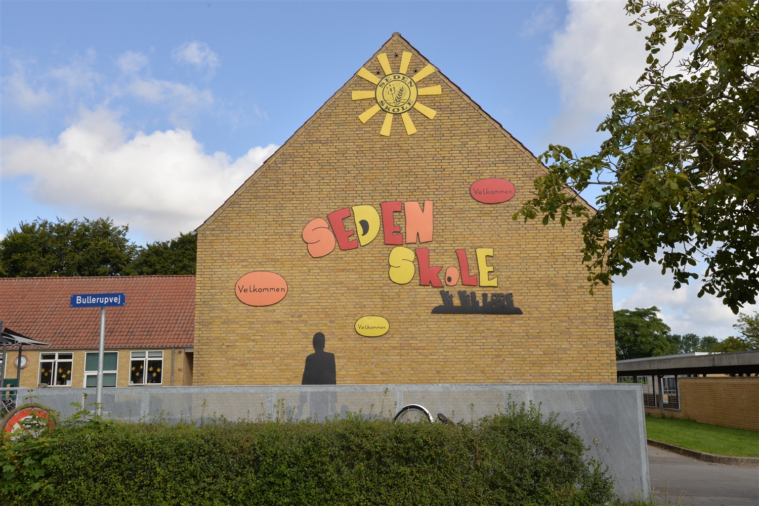 Murstensbygning med farverige skilte, hvorpå der står "Seden Skole" og "Velkommen" samt et sort skilt hvorpå der står "Vi vil lære". Forrest til venstre ses et vejskilt hvor der står "Bullerupvej". Foran bygningen ses en busk. 