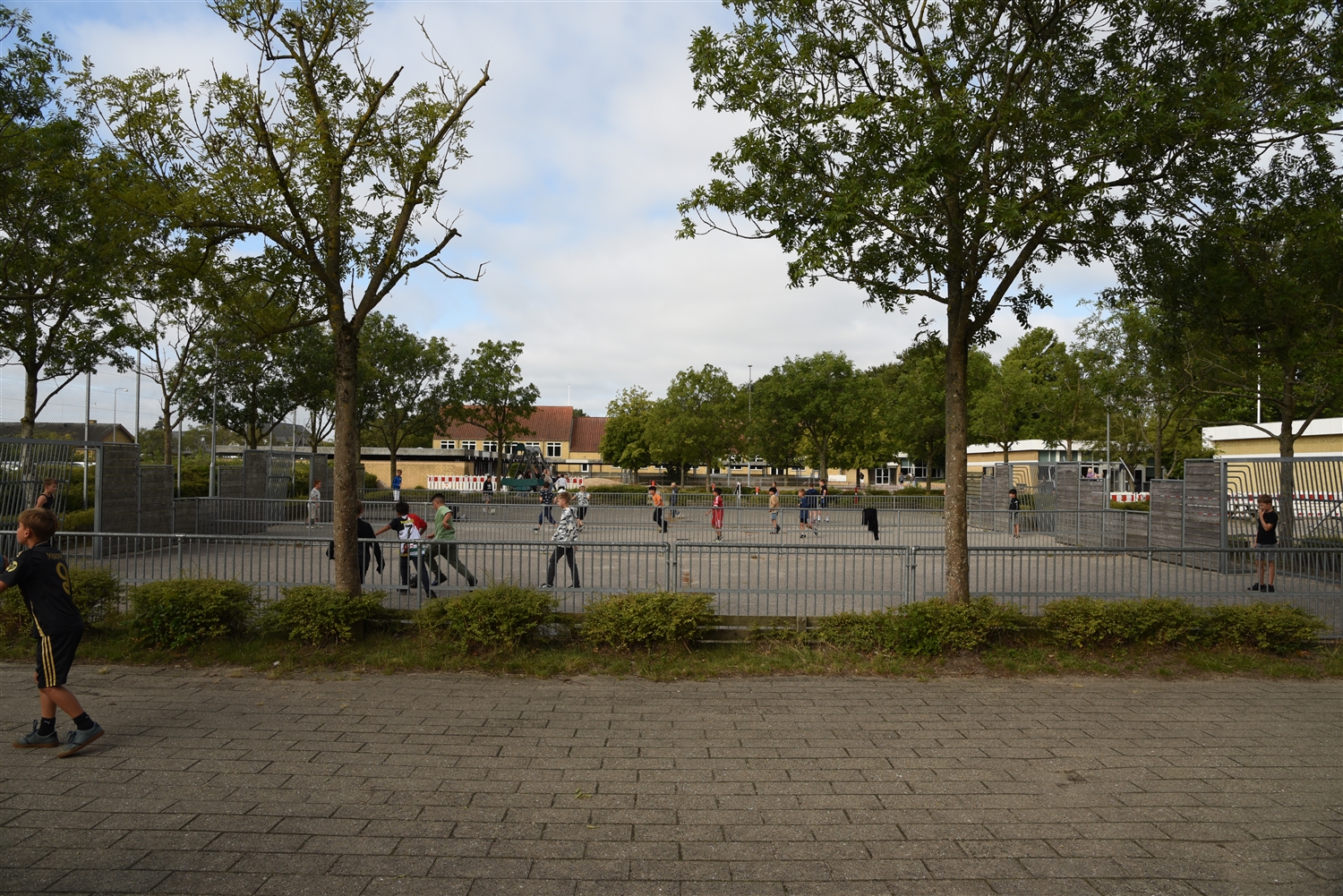 Asfalteret fodboldbane med metalhegn rundt om. Rundt om banen er der træer og buske. I baggrunden ses der bygninger. På boldbanen er der børn som spiller fodbold. 
