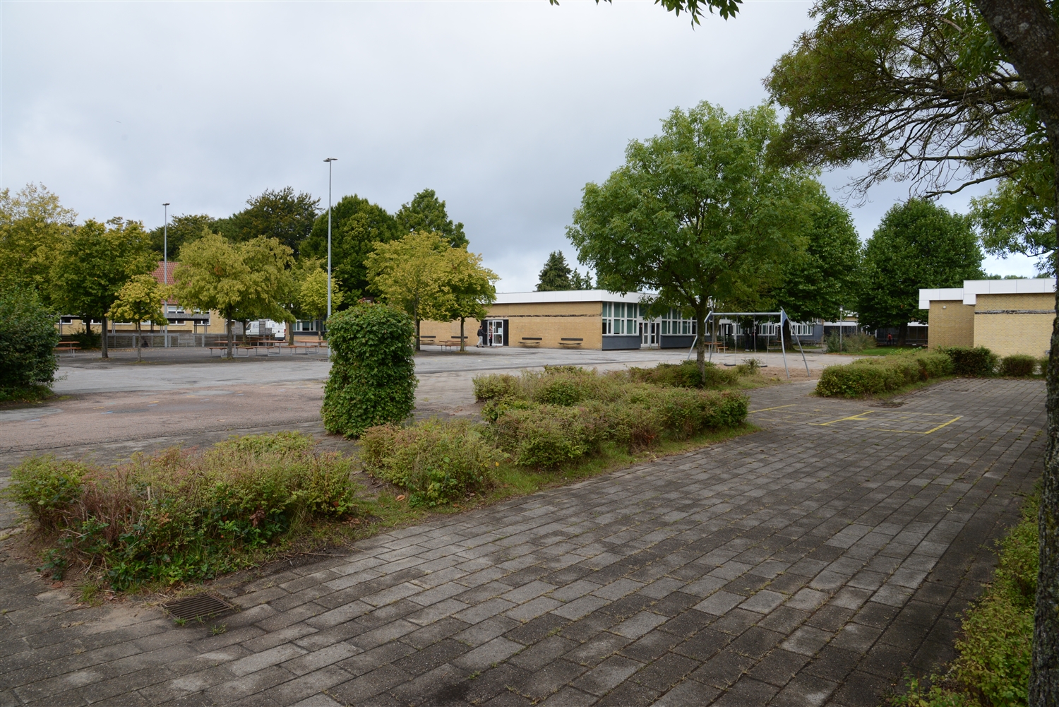 Et billede af skolebygningen på lang afstand. Foran ses træer og buske, murstensgangsti, et gyngestativ og lygtepæle.  