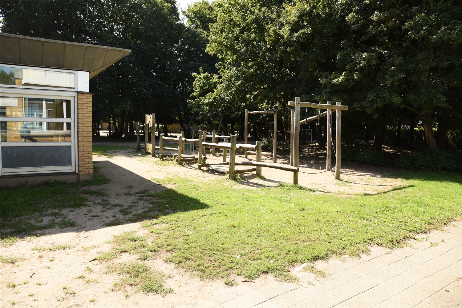 En klatrelegeplads hvor der i baggrunden ses træer. Til venstre ses lidt af en bygning med vinduer. Foran er der græs samt en murstensgangsti. 
